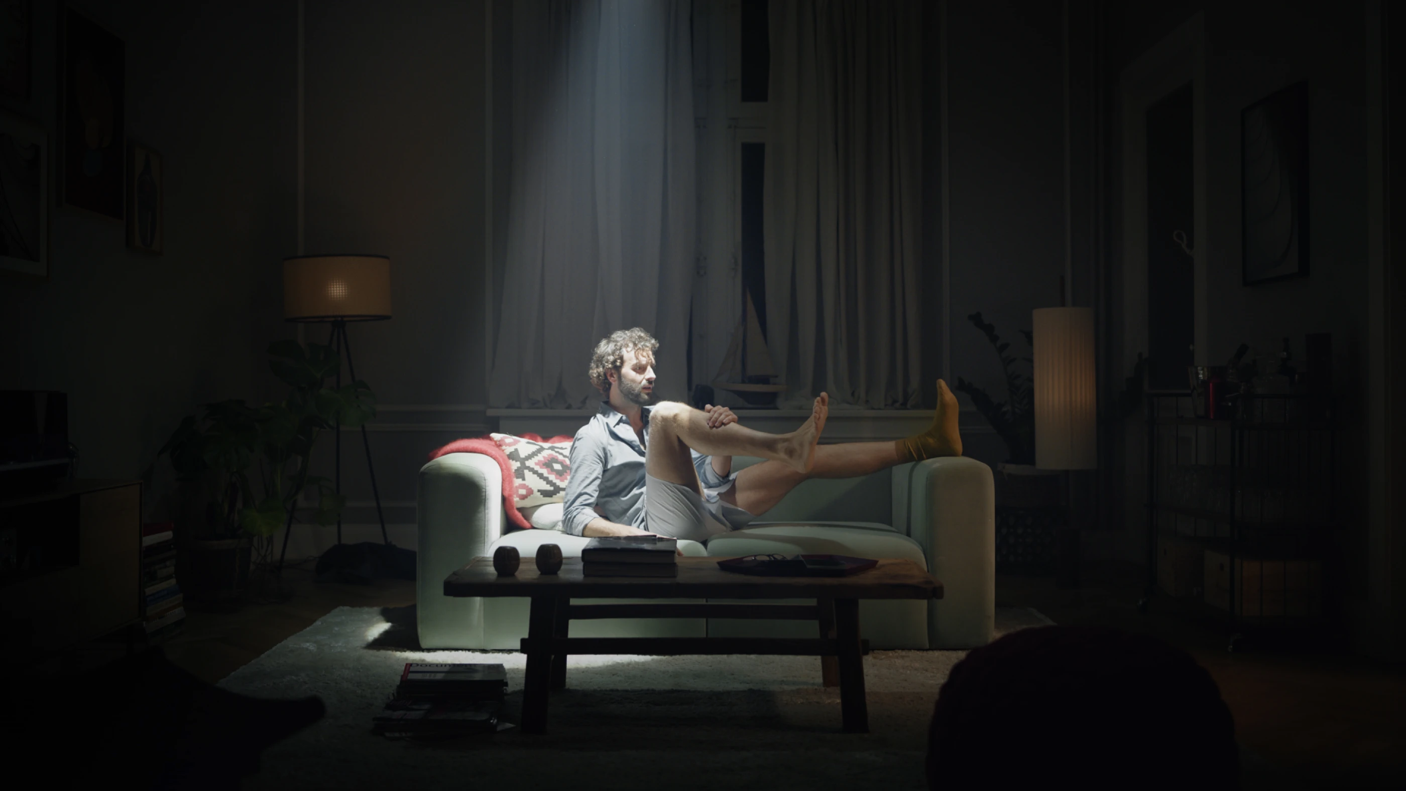 Ein Mann sitzt in einem schwach beleuchteten Wohnzimmer auf einem Sofa. Er trägt lockere Kleidung und hat die Beine auf das Sofa gelegt. Ein Lichtstrahl fällt auf ihn, während im Hintergrund Pflanzen und Lampen zu sehen sind.