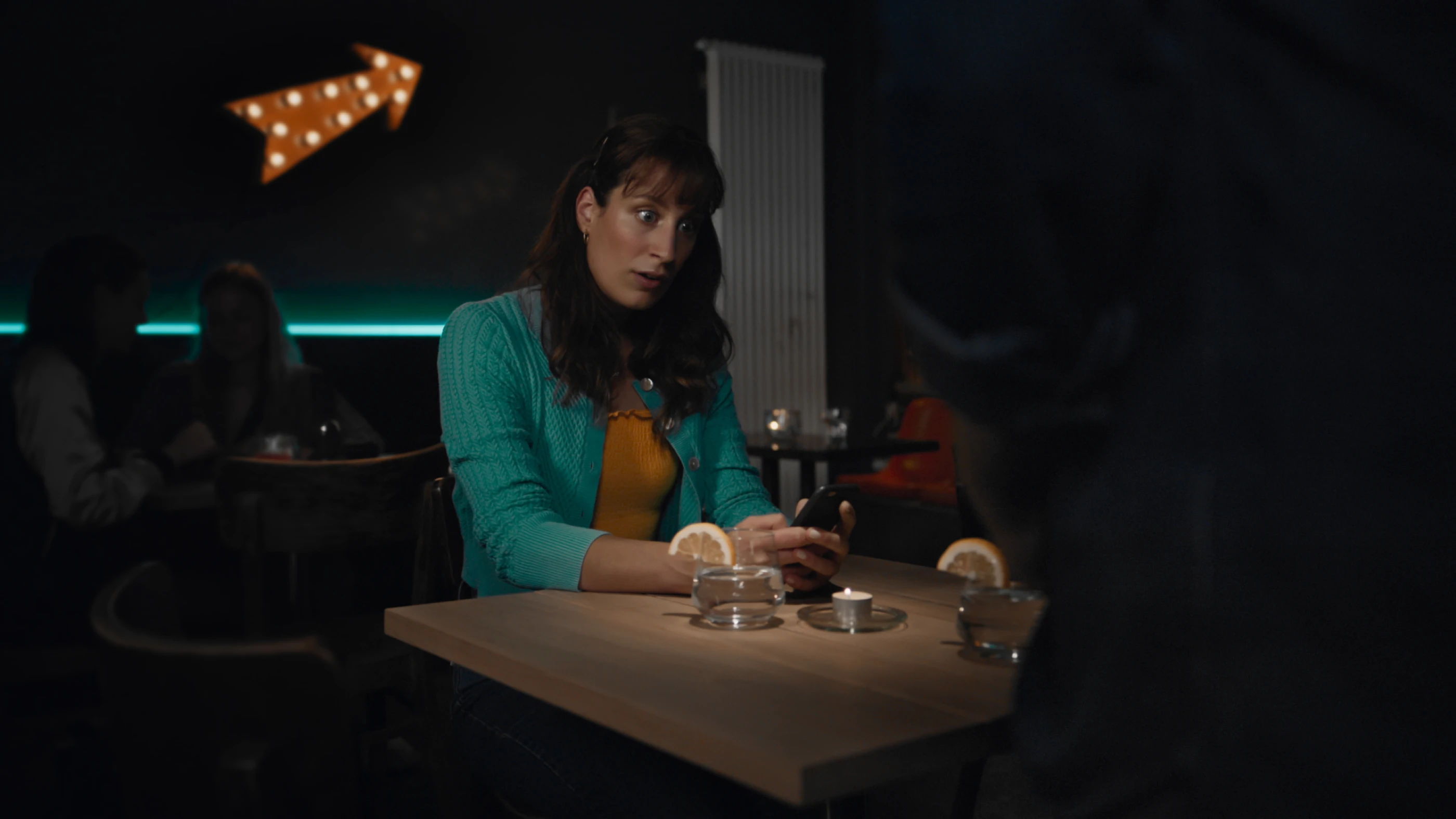 Eine Frau mit langen, dunklen Haaren sitzt an einem Tisch in einem dunklen Raum und schaut überrascht auf ihr Handy. Auf dem Tisch stehen Kerzen und zwei mit Orange dekorierte Gläser, während im Hintergrund eine beleuchtete Pfeil-S