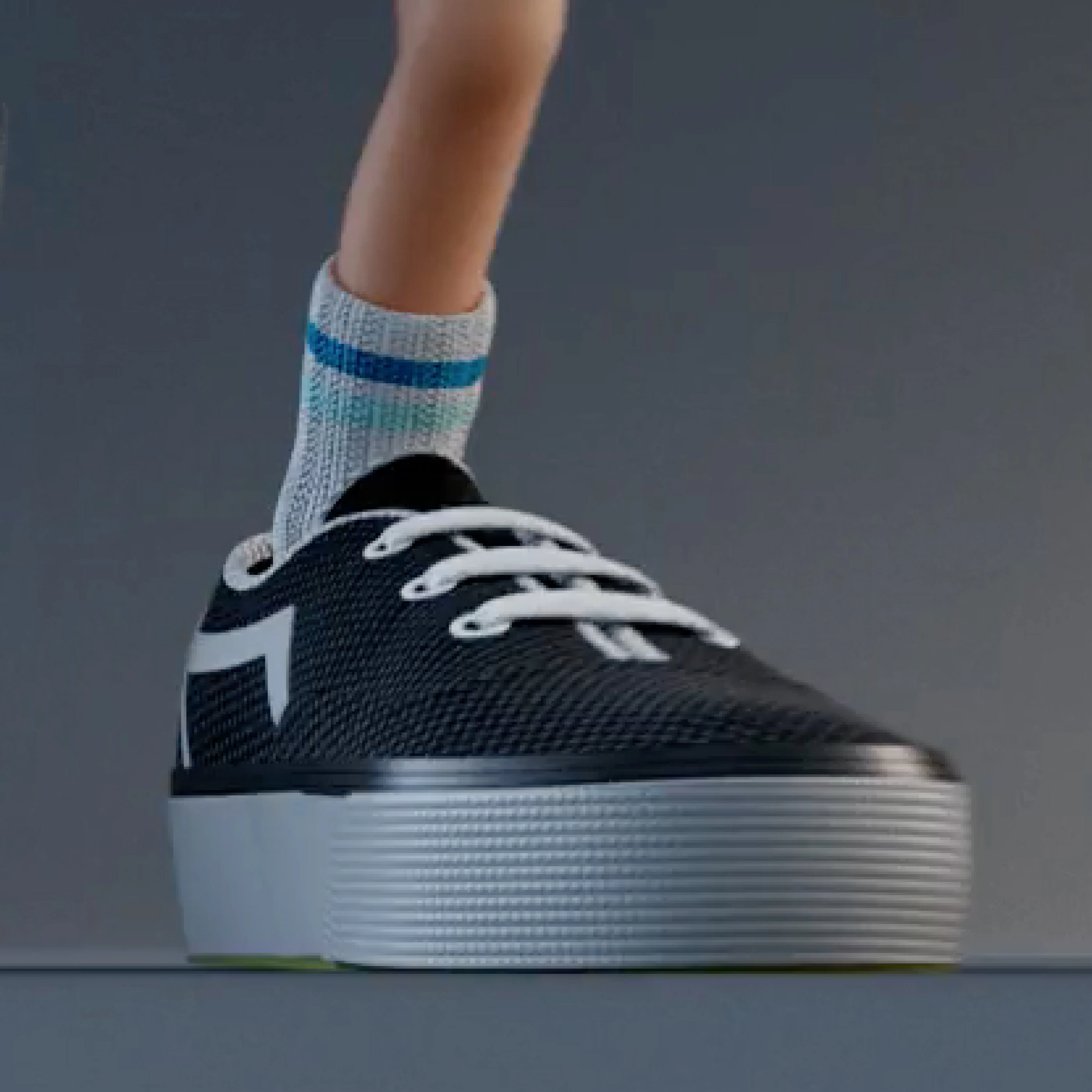 Ein großer schwarzer Sportschuh mit weißen Schnürsenkeln und einer weißen Sohle. Teil eines menschlichen Beins mit einem gestreiften Socke, steht in einer neutralen Umgebung.