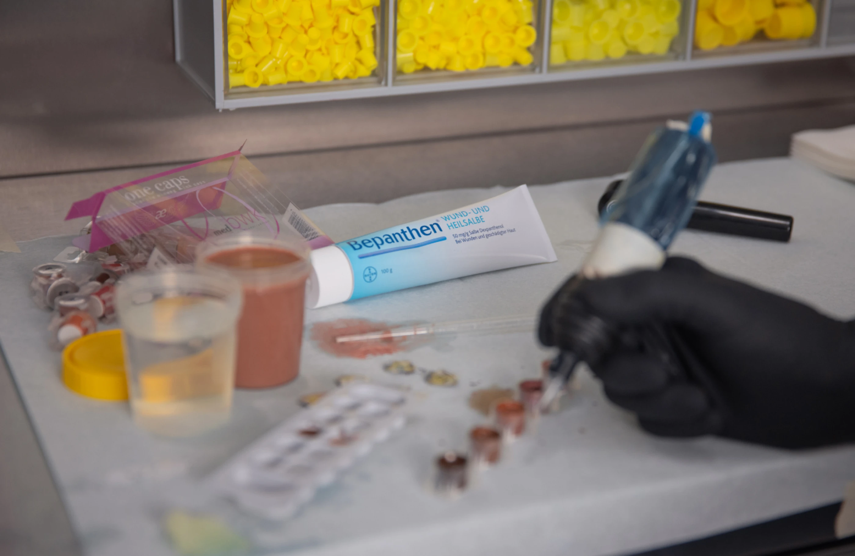 Ein Hand mit schwarzer Handschuh, die einen Tattoo- oder Permanent-Make-up-Apparat hält. Auf dem Tisch liegen ein Becher mit Flüssigkeit, eine Tube Bepanthen und verschiedene kleine Farbtöpfe. Im Hintergrund sind
