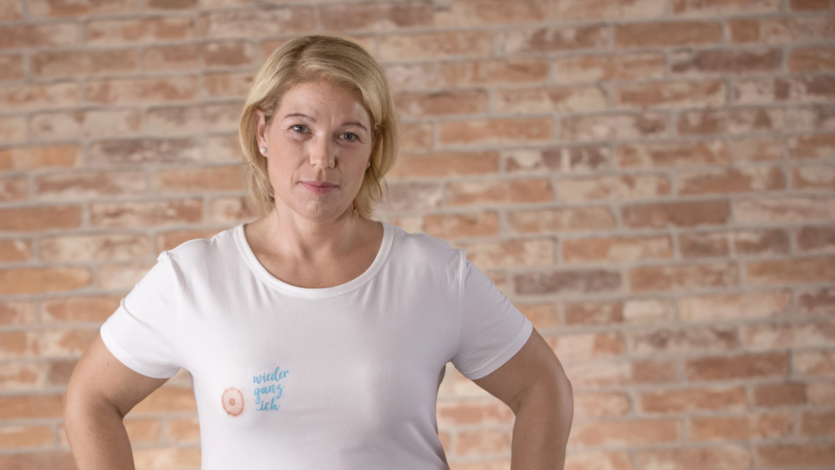 Eine Frau mit blonden Haaren trägt ein weißes T-Shirt mit einem Aufdruck. Sie steht vor einer Mauer aus Ziegelsteinen und schaut direkt in die Kamera.