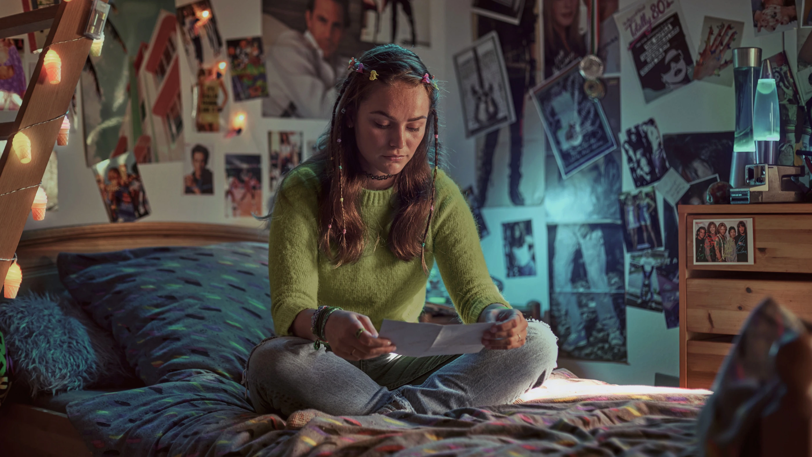 Ein Mädchen sitzt auf ihrem Bett in einem Zimmer, das mit Fotos und Postern an den Wänden dekoriert ist. Sie trägt einen grünen Pullover und schaut nachdenklich auf ein Foto, das sie in den Händen hält. Warmes Licht