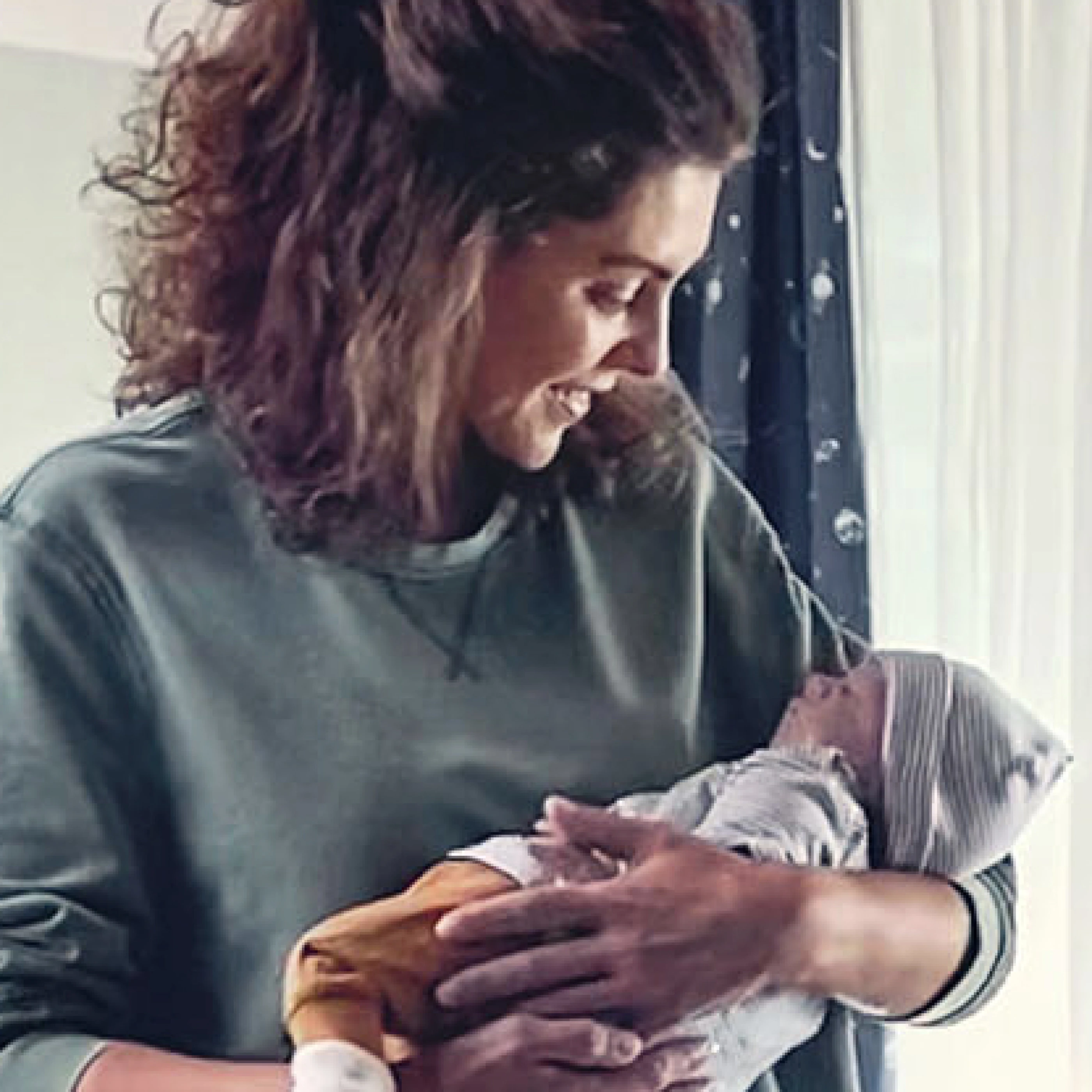 Eine lächelnde Frau hält ein Neugeborenes in ihren Armen. Das Baby trägt einen gestreiften Hut und einen bunten Overall. Die Szene strahlt Wärme und Zuneigung aus.