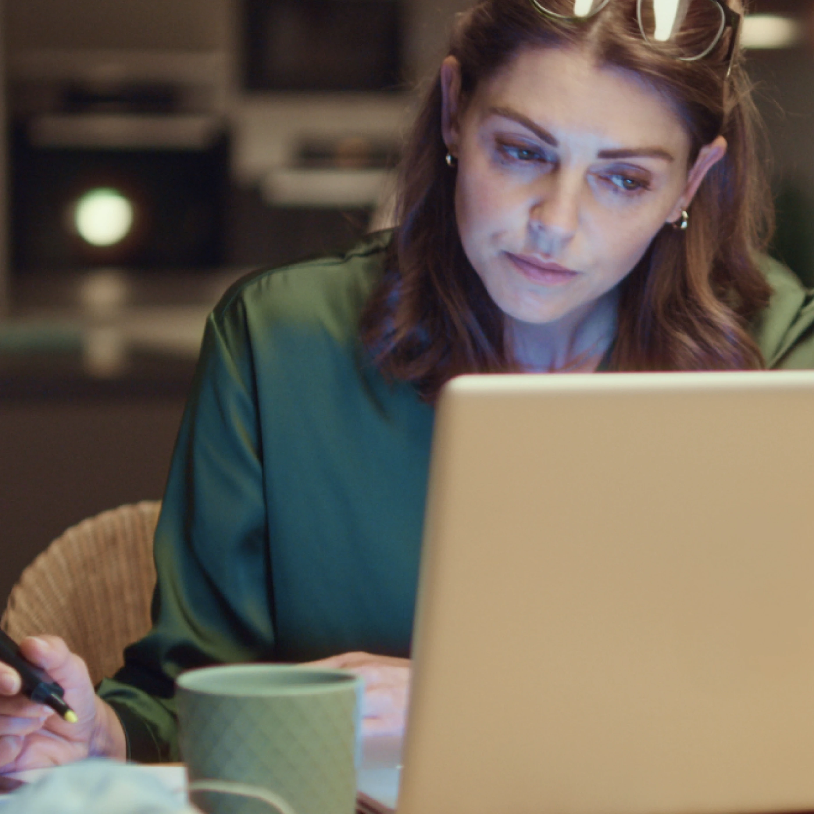 Eine Frau in einem grünen Oberteil sitzt an einem Tisch und arbeitet konzentriert an einem Laptop. Sie hält einen Stift in der Hand und hat eine Tasse neben sich. Hinter ihr ist ein moderner Raum mit gedämpftem Licht zu