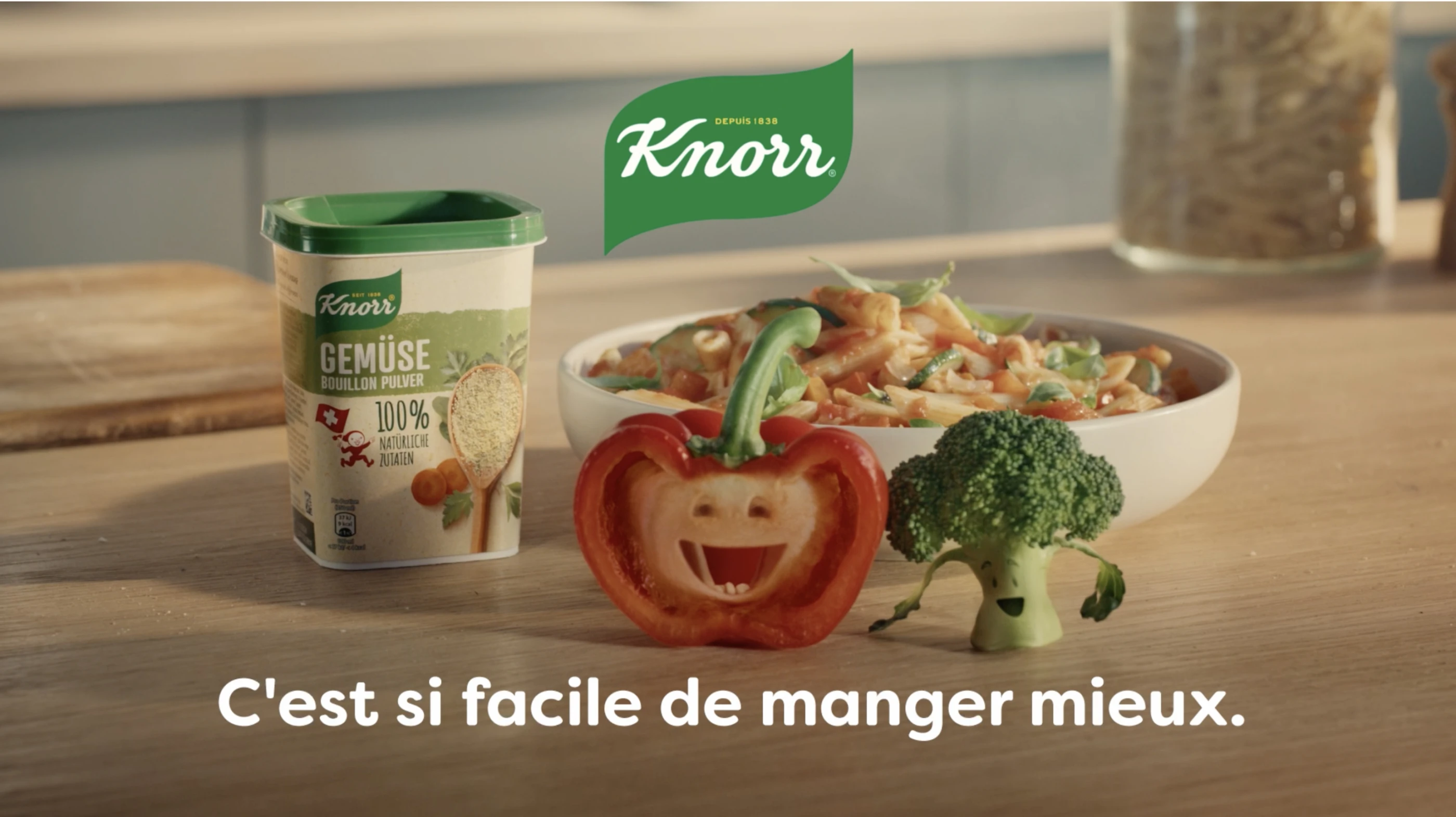 Eine Packung Knorr Gemüse-Bouillon-Pulver steht neben einer Schüssel bunter Gemüsenudeln auf einem Tisch. Davor steht eine angeschnittene Paprika und ein Brokkoli-Röschen, die Gesichter haben und fröhlich singen. Darunter steht der französische Text: "C'est si facile de manger mieux.".