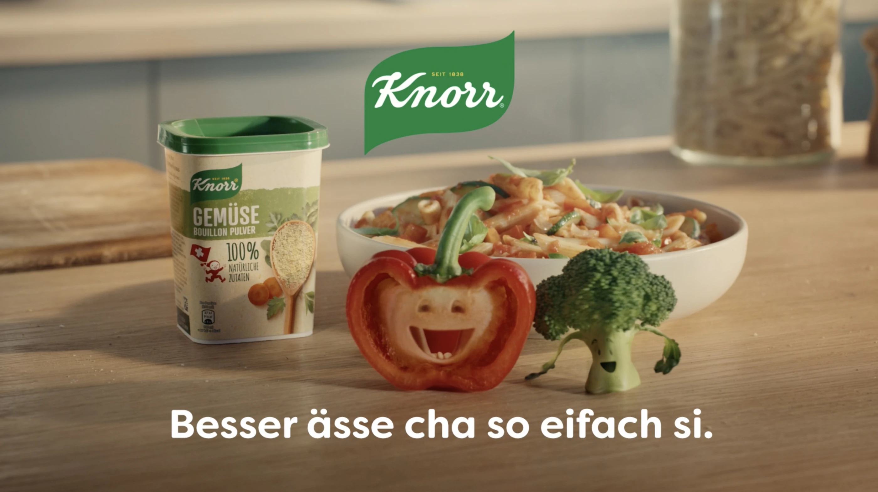 Eine Packung Knorr Gemüse-Bouillon-Pulver steht neben einer Schüssel bunter Gemüsenudeln auf einem Tisch. Davor steht eine angeschnittene Paprika und ein Brokkoli-Röschen, die Gesichter haben und fröhlich singen. Darunter steht ein Text in Schweizerdeutsch: "Besser ässe cha so eifach si.".