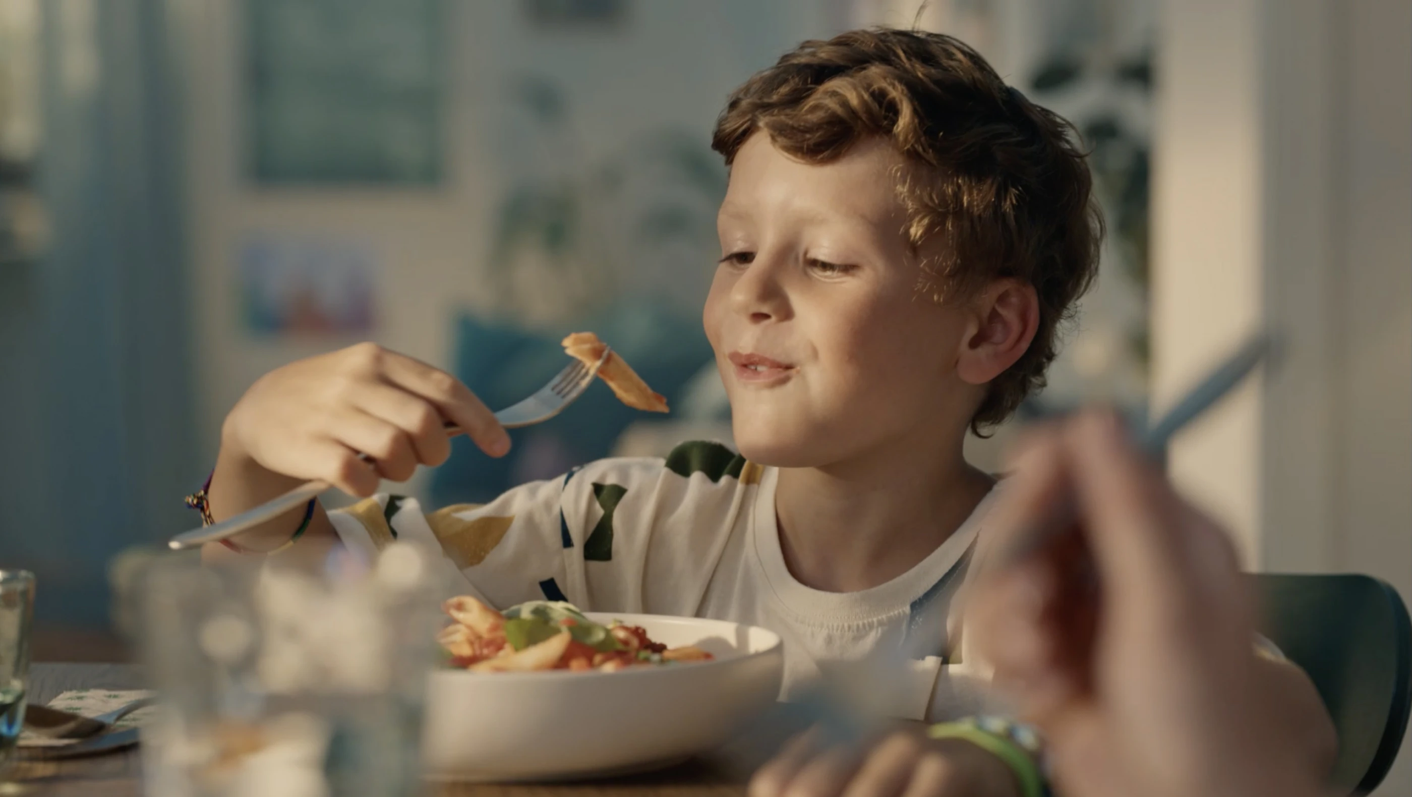 Ein junger Junge mit lockigem Haar sitzt am Tisch und genießt sein Essen. Er zeigt ein zufriedenes Lächeln, während er mit einer Gabel Pasta aufnimmt. Der Tisch ist mit weiteren Geschirrteilen und Gläsern gedeckt.