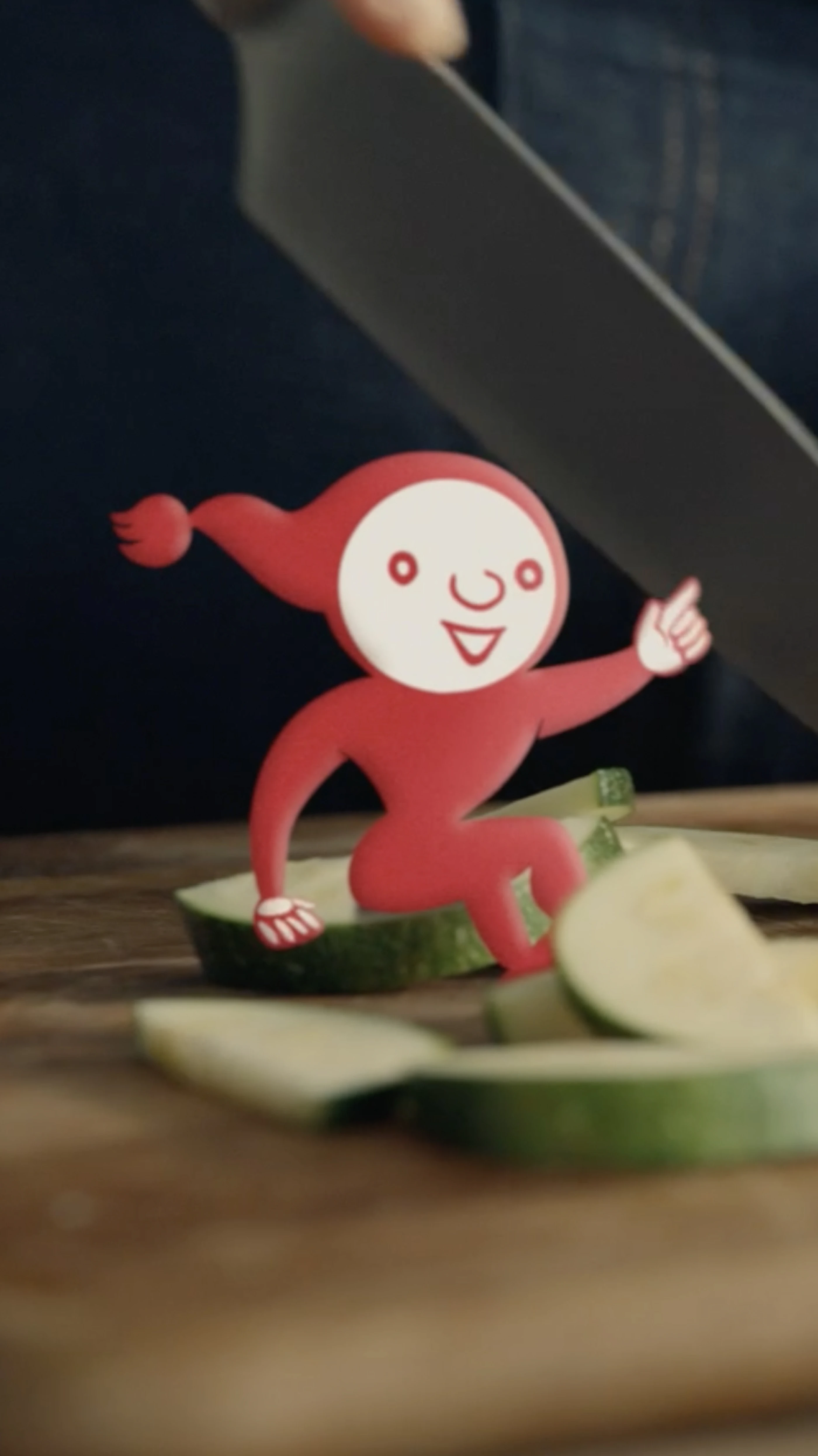 Der fröhlich, rot Cartoon-Charakter Knorrli mit einem breiten Lächeln sitzt auf geschnittenen Zucchinistücken und hebt einen Finger nach oben. Im Hintergrund ist ein Messer sichtbar.