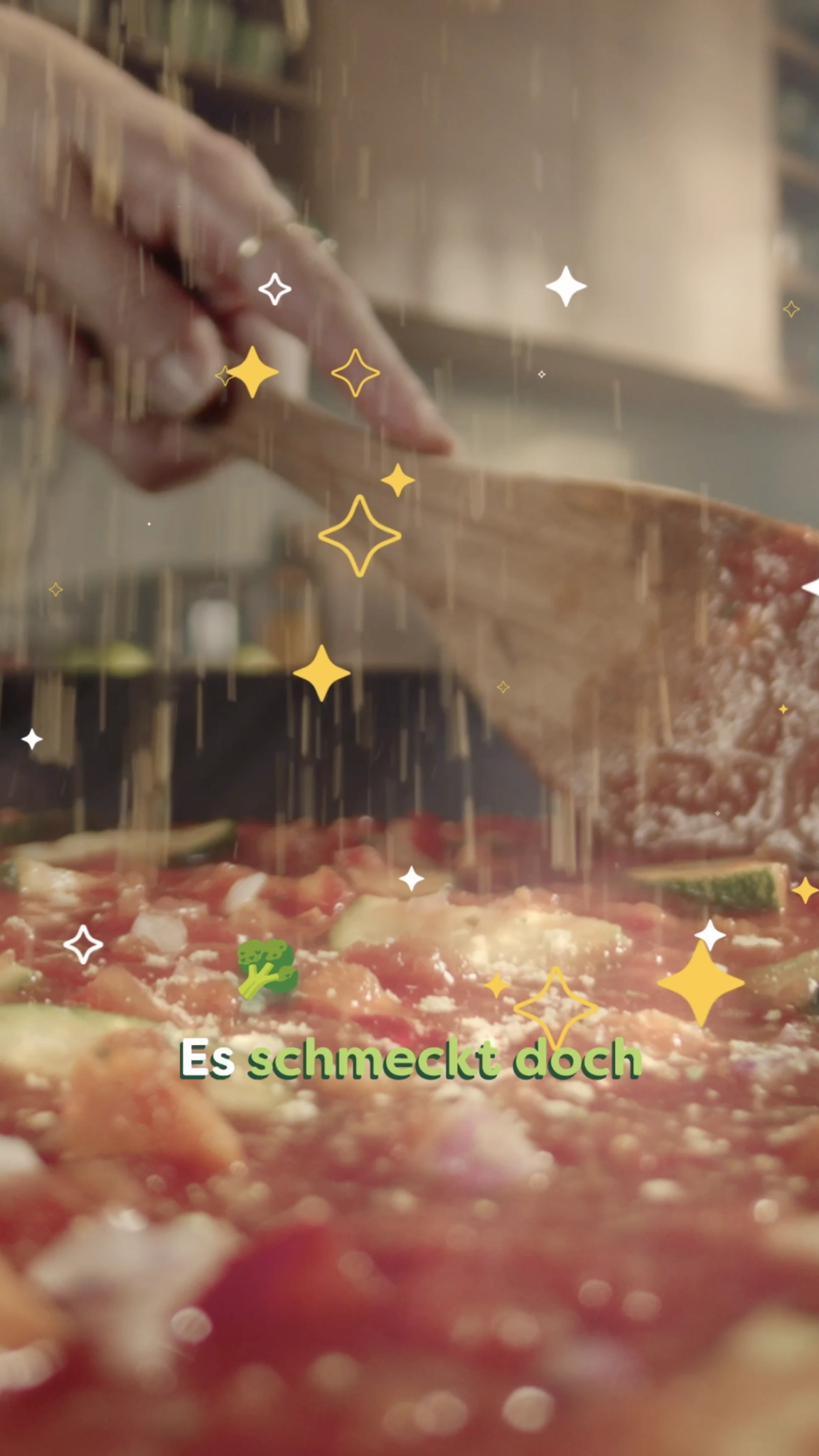 Eine Hand rührt mit einem Holzlöffel durch eine eine frisch zubereitete Gemüsesuppe. Parmesan fällt von oben in die Suppe. Illustrierte Sterne und der Spruch "Es schmeckt doch" verleihen der Szene einen zauberhaften Touch.