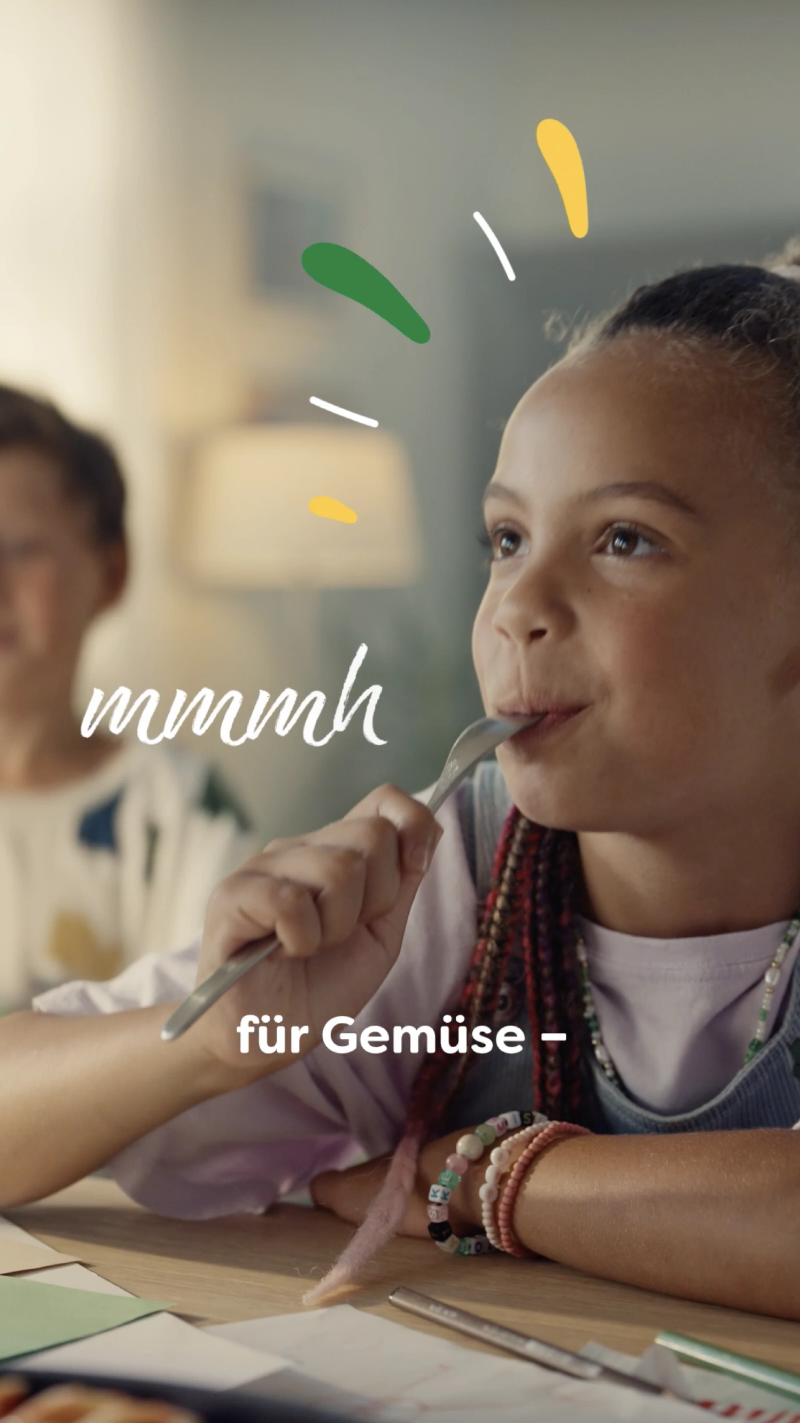Ein Mädchen mit Zöpfen genießt einen Bissen und blickt zufrieden. Im Hintergrund ist ein weiterer Junge sichtbar. Der Text im Vordergrund lautet "mmmh - für Gemüse"