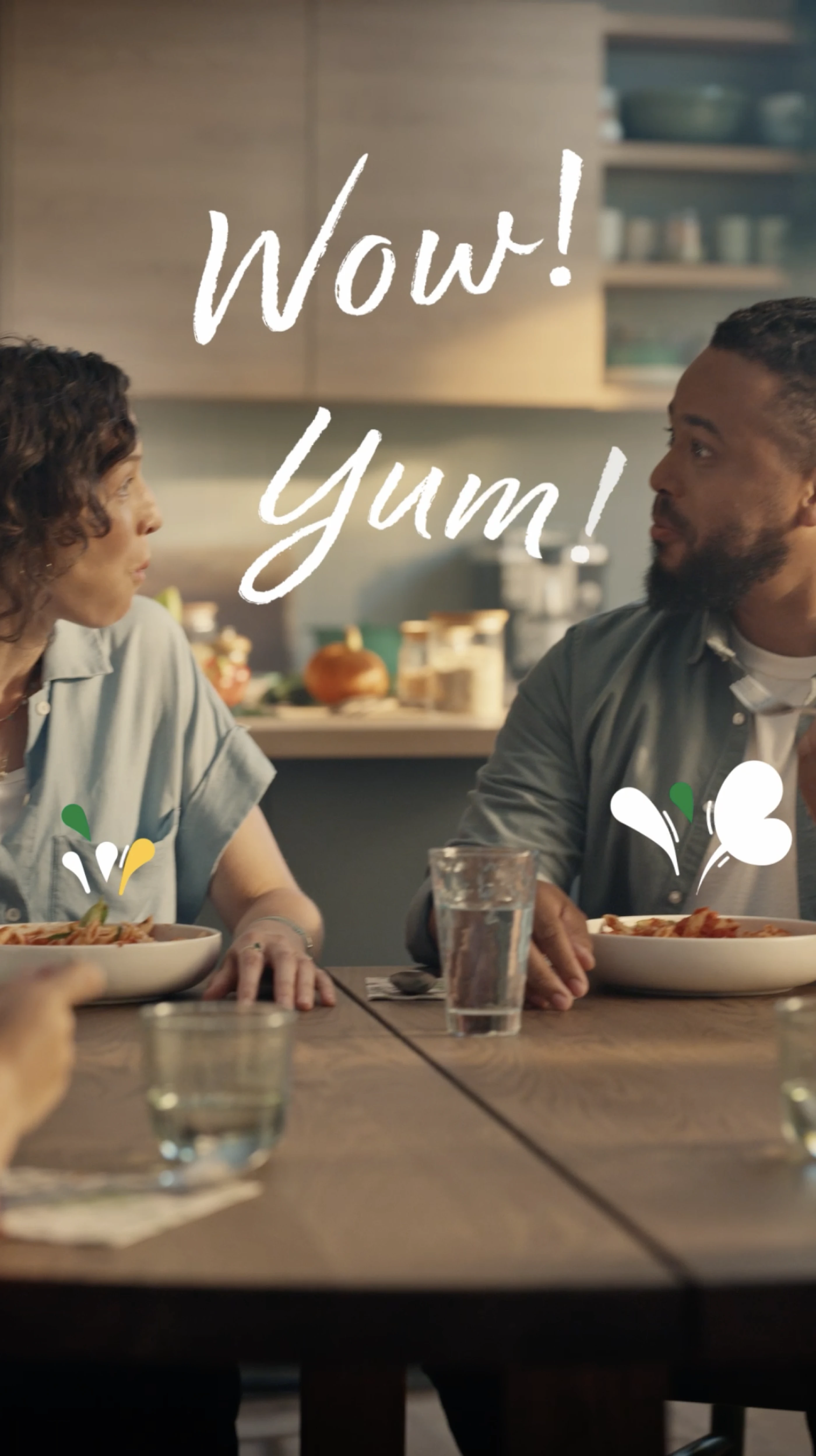 Ein Paar sitzt an einem Tisch und schaut sich überrascht an, während sie eine Mahlzeit genießen. Vor ihnen stehen zwei Schalen mit Essen und Gläser mit Wasser. Das Wort "Wow!" und "Yum!" sind kreativ über das Bild verteilt.