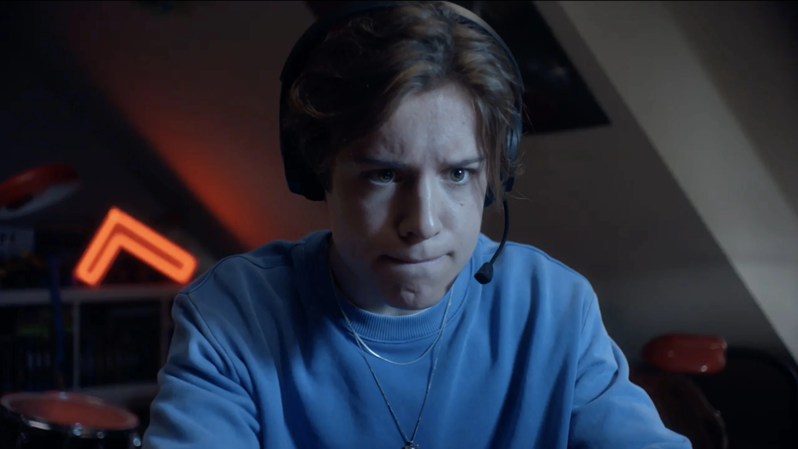 Ein junger Mann mit Kopfhörern schaut konzentriert auf einen Bildschirm. Er trägt ein hellblaues Sweatshirt und hat eine ernsthafte Mimik. Im Hintergrund ist eine rote Neon-Beleuchtung zu sehen.