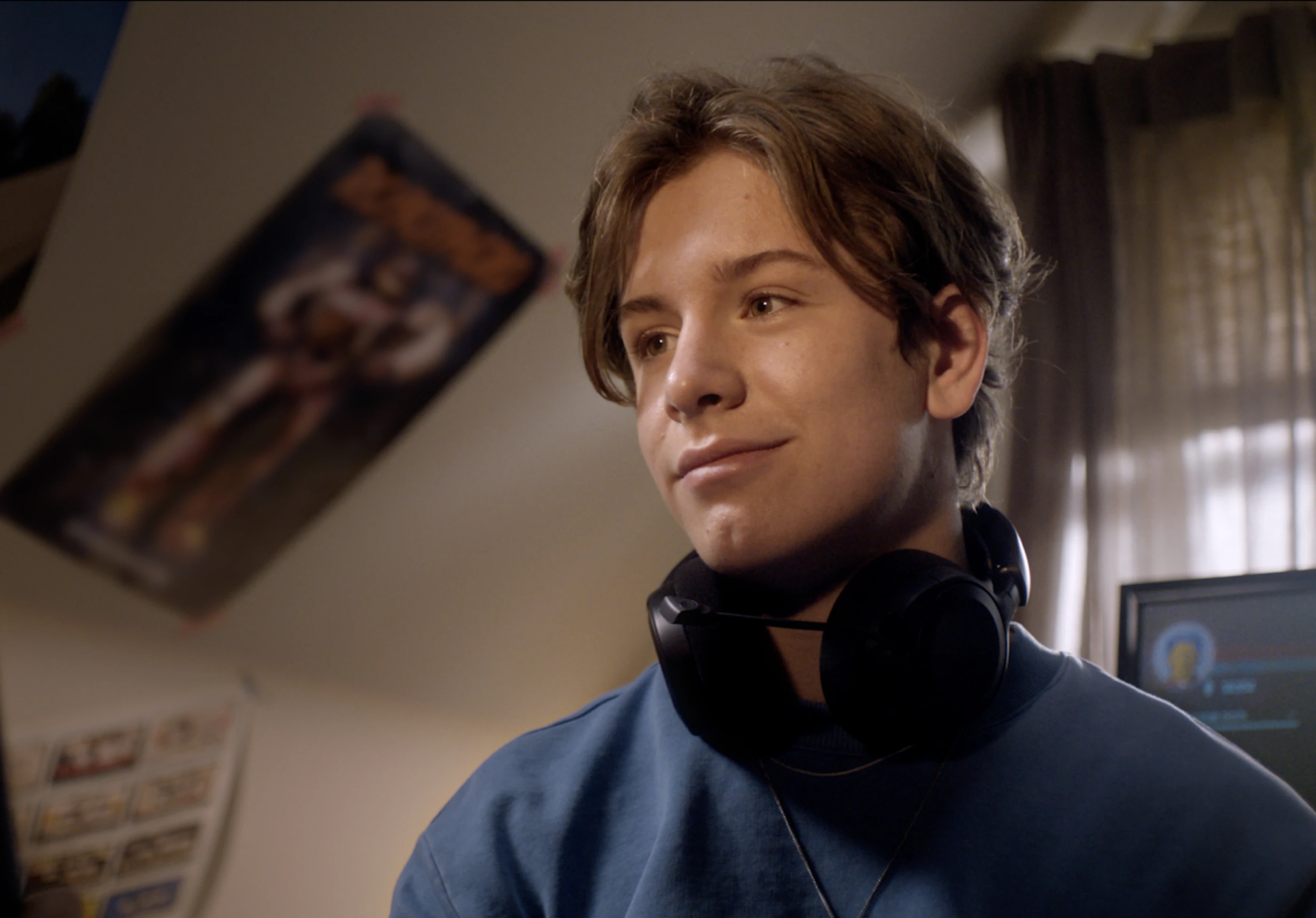 Ein junger Mann mit Kopfhörern trägt ein blaues Sweatshirt. Er lächelt leicht und schaut nachdenklich. Im Hintergrund sind Poster und ein Computer sichtbar.