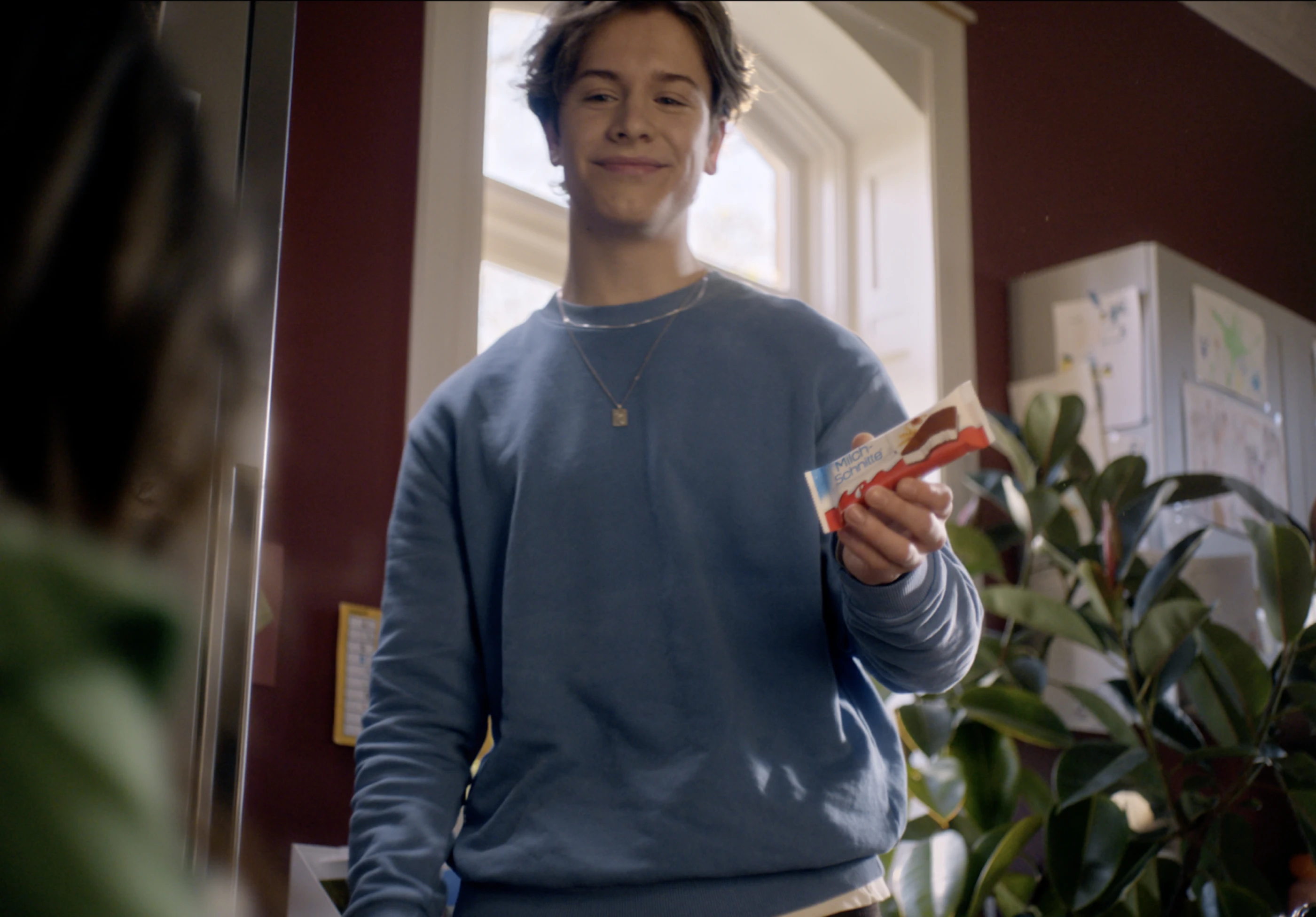 Ein junger Mann hält eine Milchschnitte in der Hand und lächelt freundlich. Im Hintergrund ist eine Pflanze und ein Wandschrank mit Zeichnungen zu sehen.