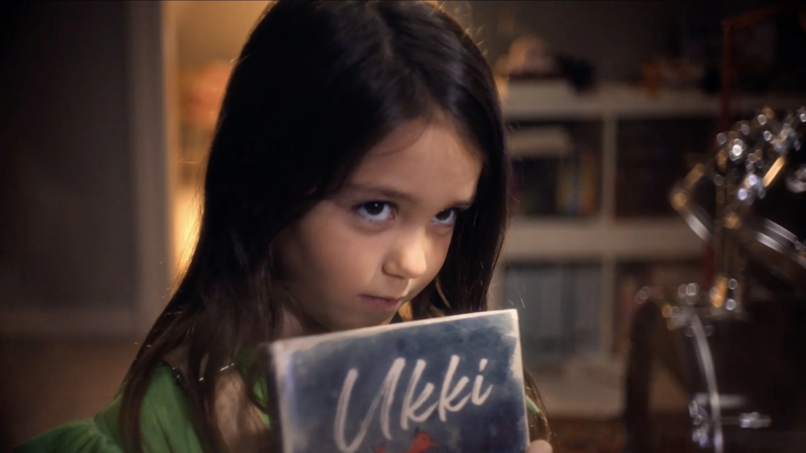 Ein Mädchen mit langen, dunklen Haaren hält ein Buch mit dem Titel "Ukki" in der Hand und schaut schüchtern. Im Hintergrund sind Bücherregale und eine gemütliche Atmosphäre sichtbar.