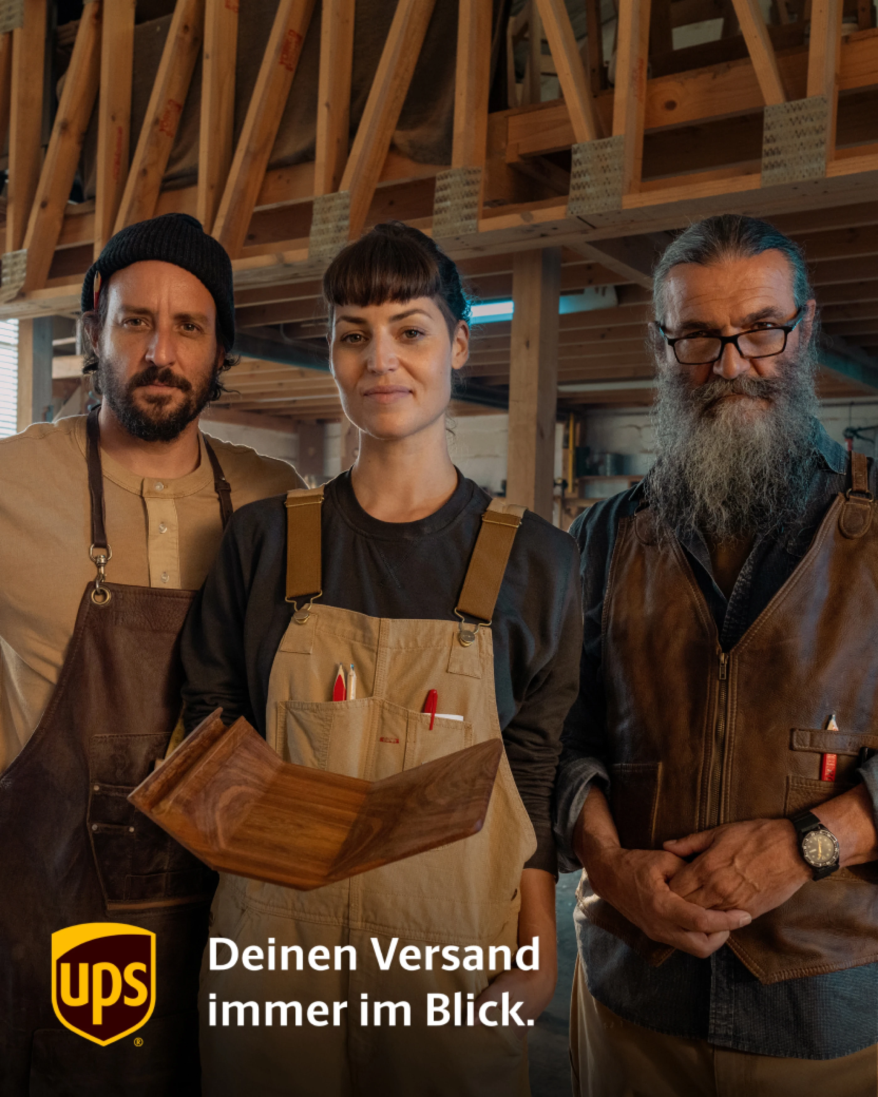 Drei Handwerker:innen in einer Werkstatt stehen zusammen, schauen in die Kamera und halten ein Holzelement. Im Hintergrund sind Holzbearbeitungswerkzeuge und Holzregale zu sehen. Das UPS-Logo und der Slogan "Deinen Versand immer im Blick" stehen unten.