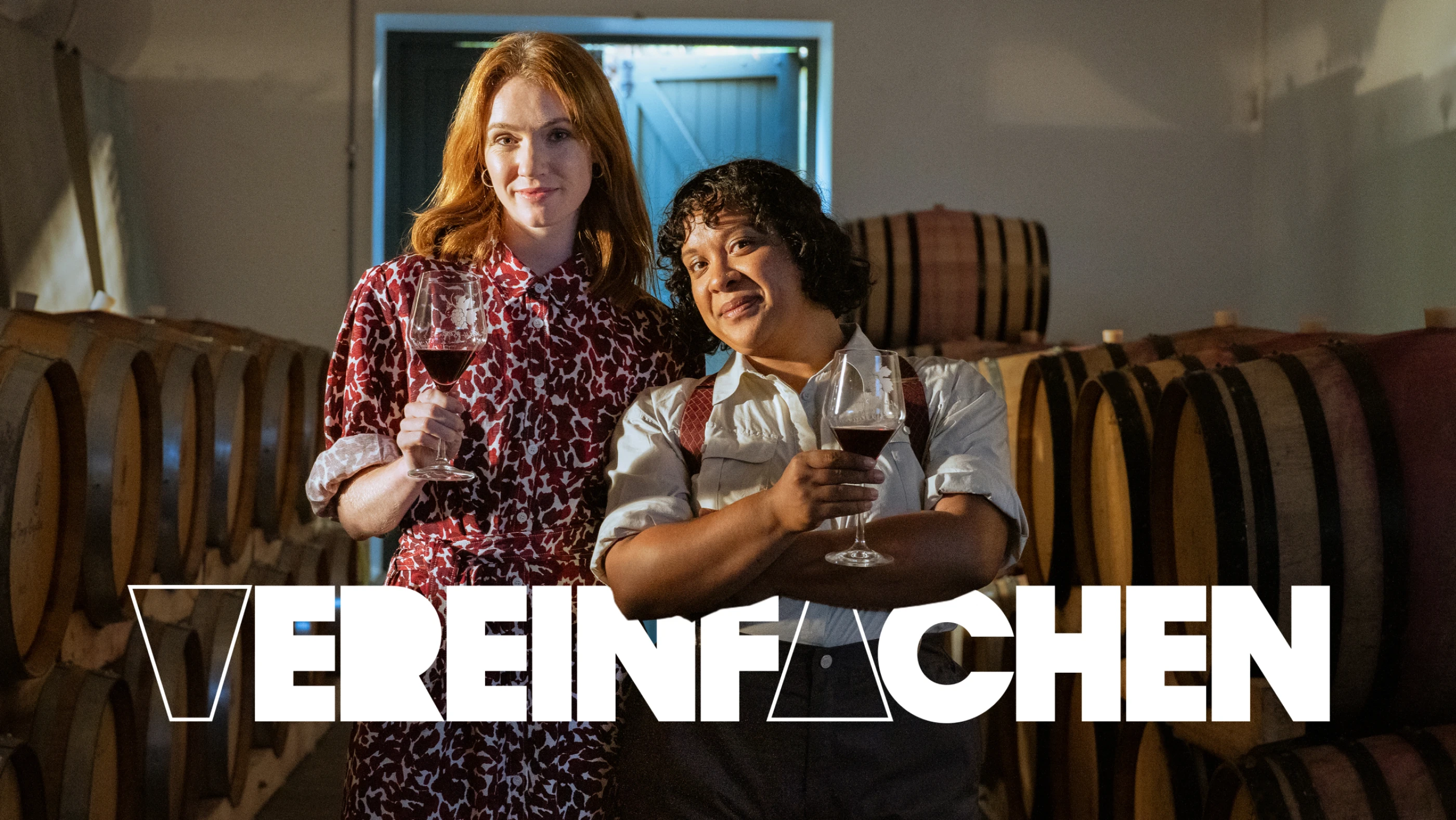 Zwei Personen stehen in einem Weinkeller, beide halten ein Glas Rotwein und schauen in die Kamera. Im Hintergrund sind Holzfässer zu sehen. Der verspielte Schriftzug "VEREINFACHEN" ist prominent platziert.